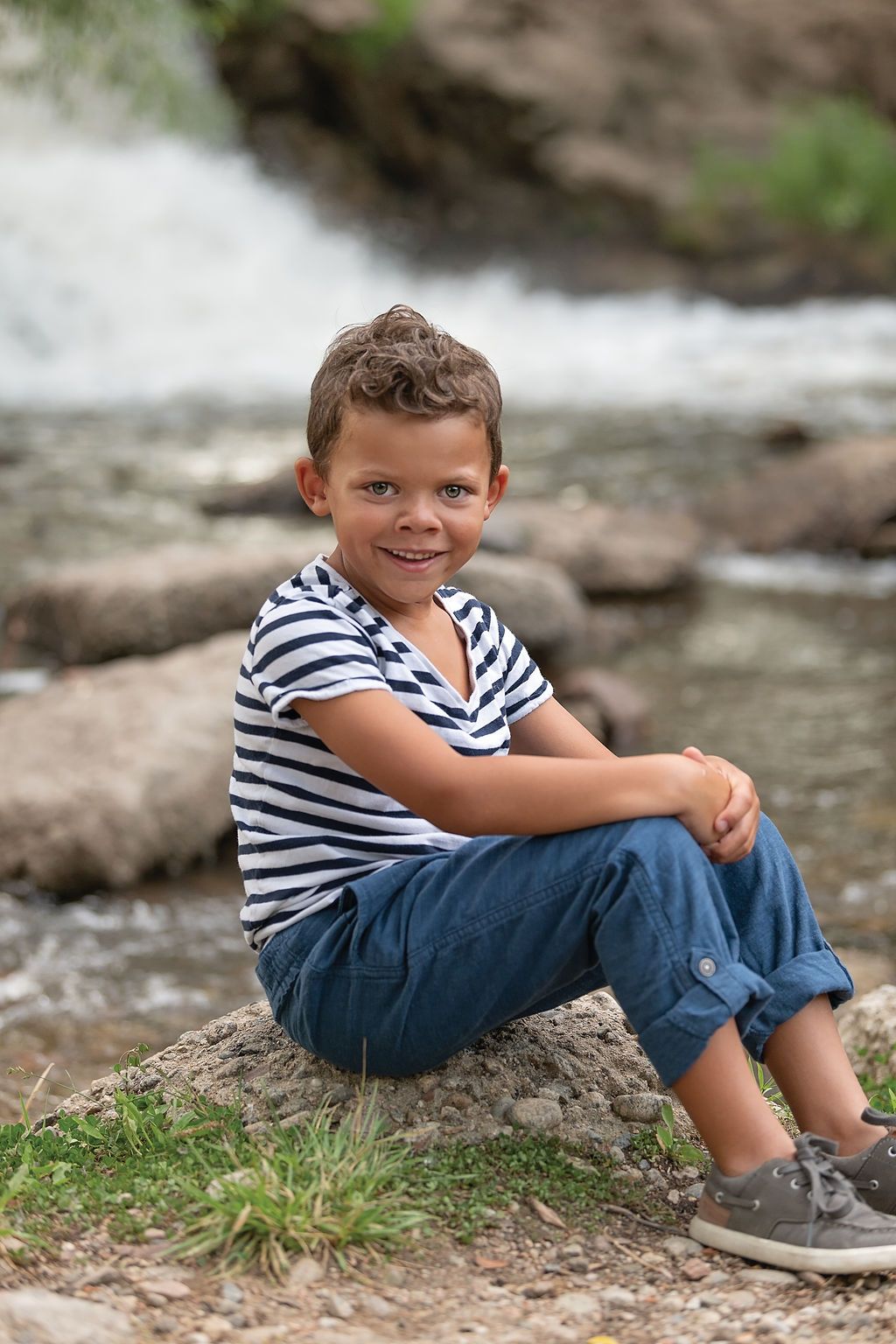 Child posing in front of a waterfall framed by vibrant fall foliage during a golden hour mini session in Longmont, Color
