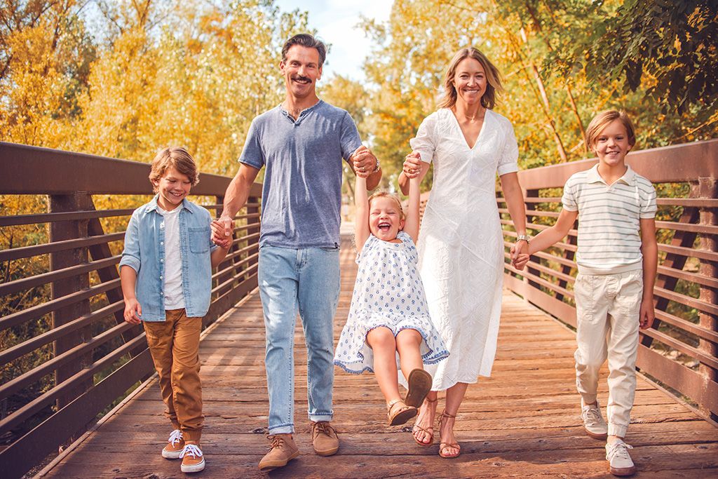 Family walking playfully swinging child on a wooden footbridge during a Longmont photography session at Golden Ponds.