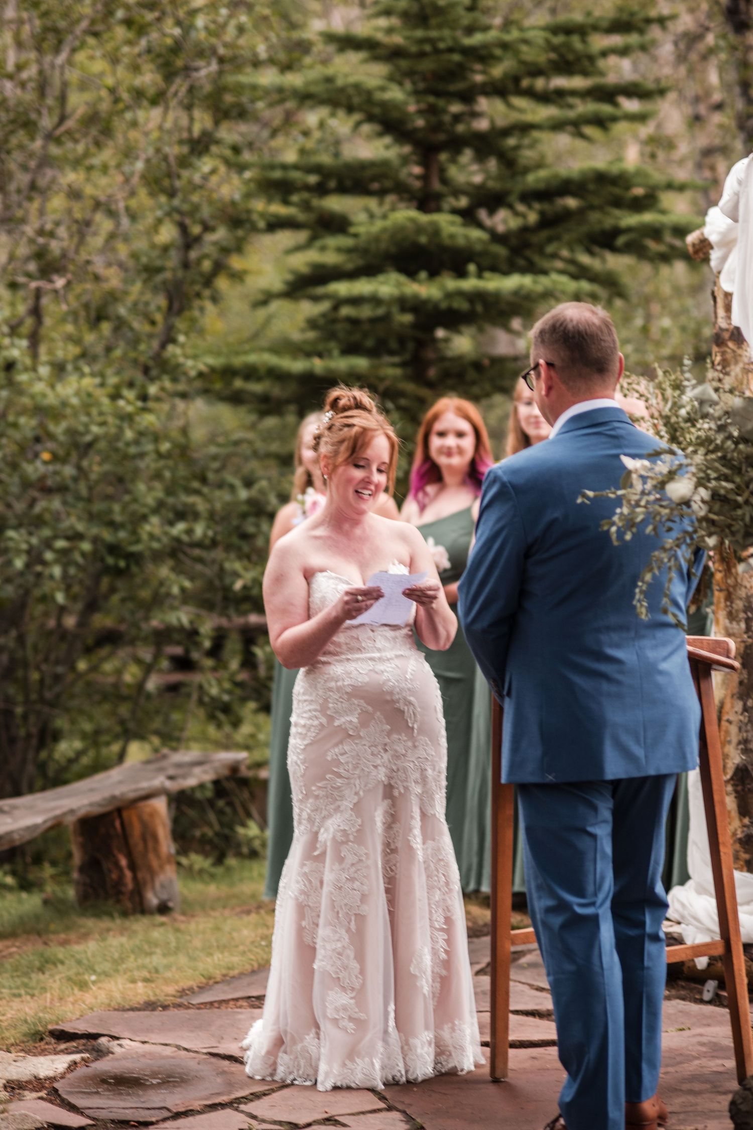 Bride in lace gown reads vows to groom in blue suit during outdoor wedding ceremony surrounded by evergreen trees. Riversong Inn Retreat