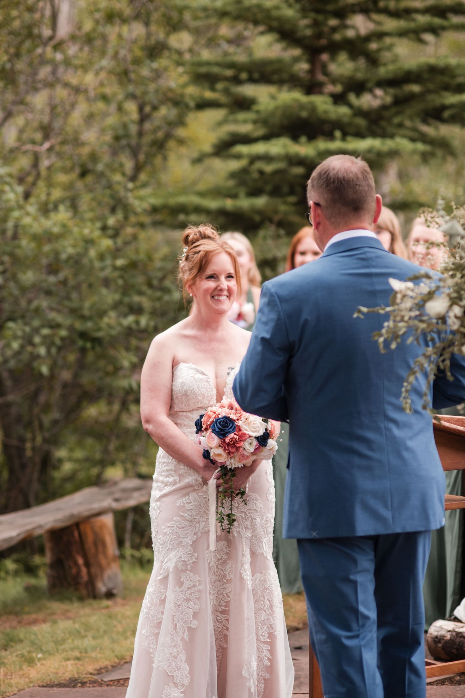 Bride in lace gown holds pink and navy bouquet, smiling at groom in blue suit during outdoor wedding ceremony. Exchange wedding vows
