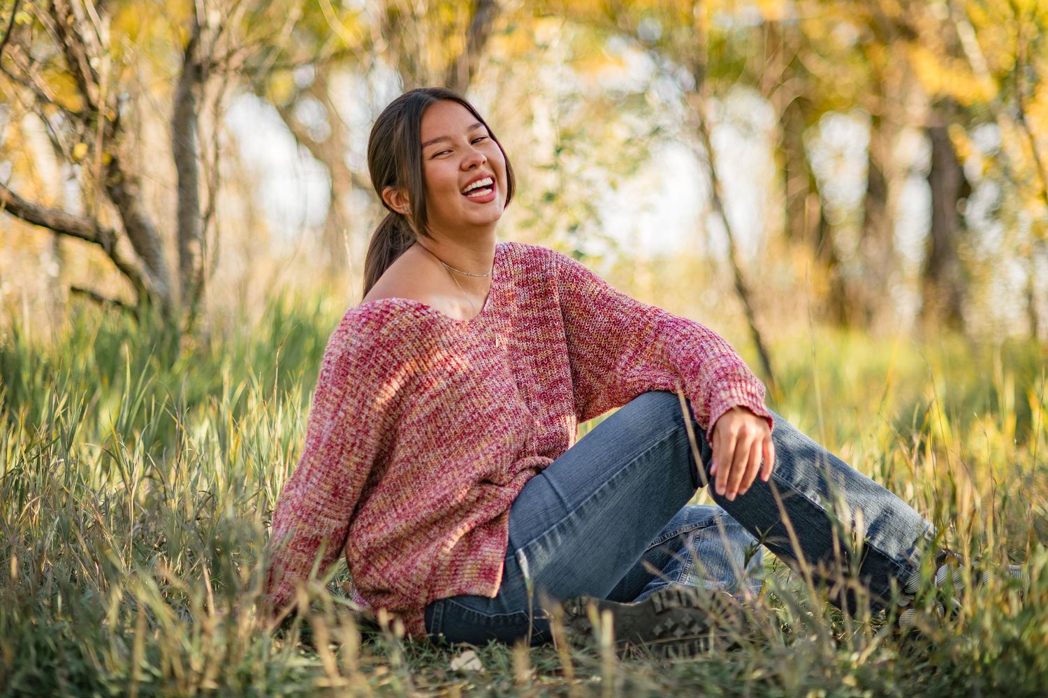 Young woman in pink sweater and jeans laughing outdoors, sitting in grass with autumn trees in background.Senior photo session at Coot Lake