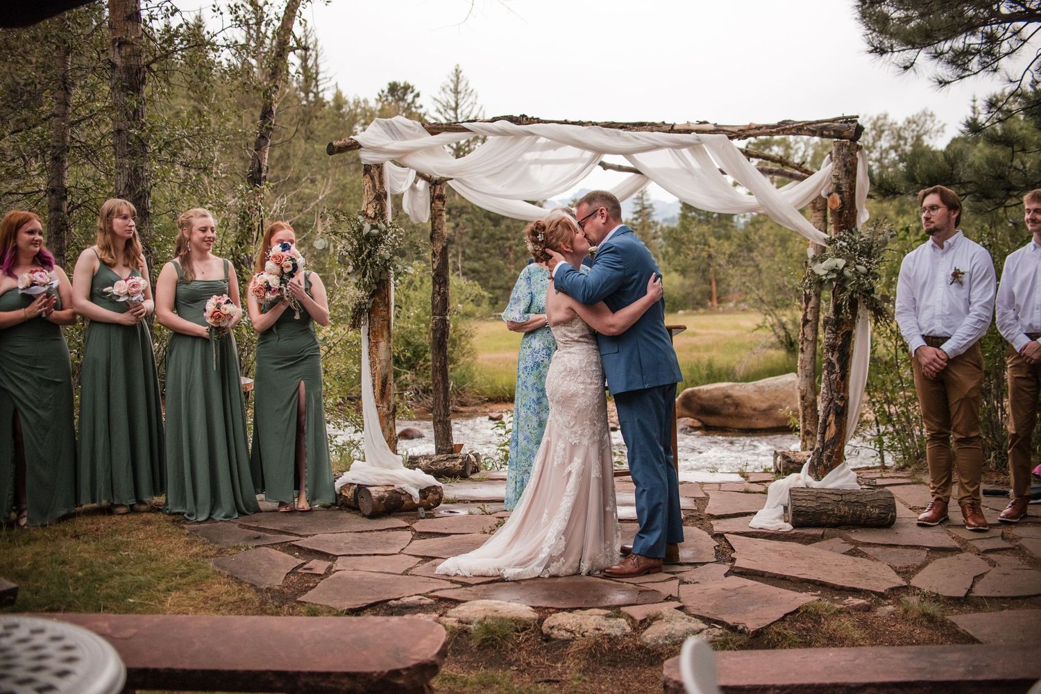 Bride and groom share first kiss under rustic wooden arch with bridesmaids in green dresses at outdoor forest wedding. Wedding Photography
