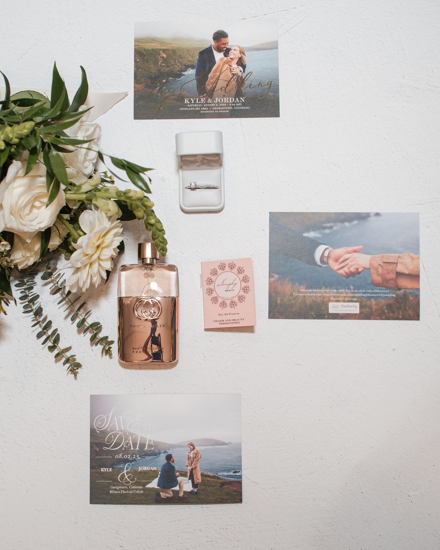 Flat lay of wedding stationery suite with save the date cards, perfume bottle, ring box, and white floral bouquet.