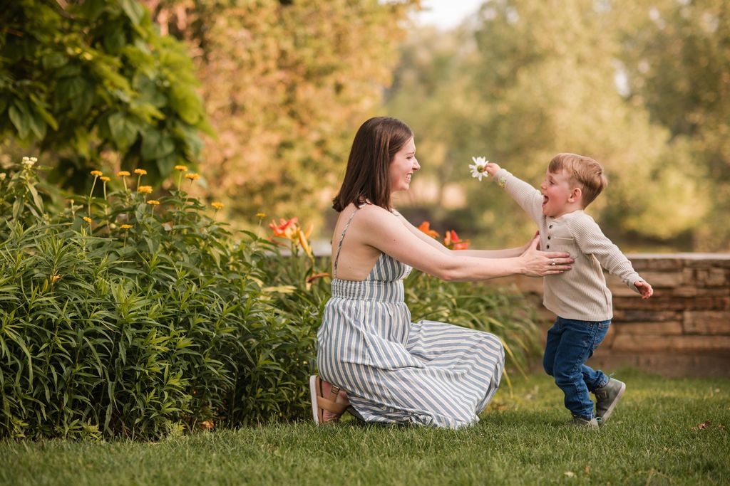 A mother kneels on green grass, smiling as her toddler son reaches toward her in a sunny garden Photography  Longmont Sandstone Ranch