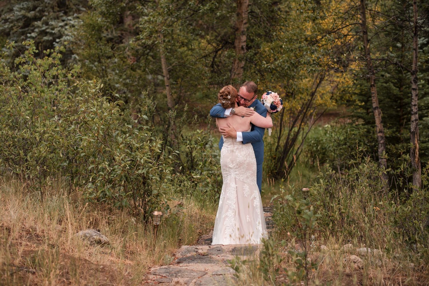 A bride and groom share a romantic embrace surrounded by lush green trees in an outdoor forest wedding setting. First Look Estes Park