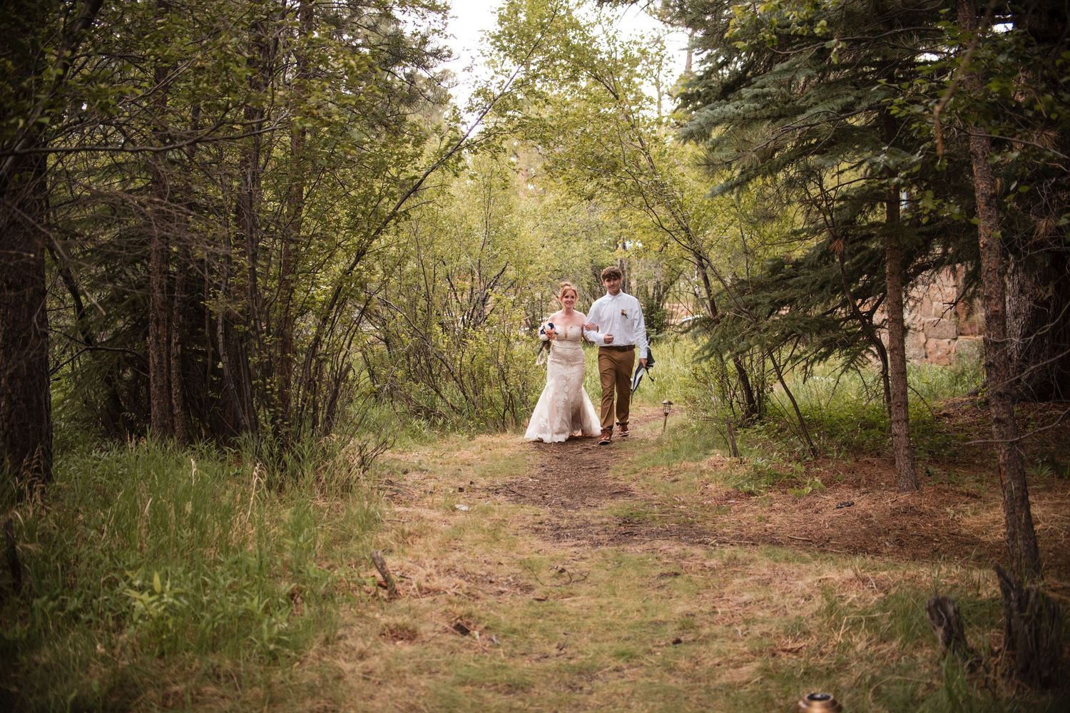 A bride and son walk hand in hand along a forest path surrounded by lush green trees on their wedding day to the altar. Estes park wedding