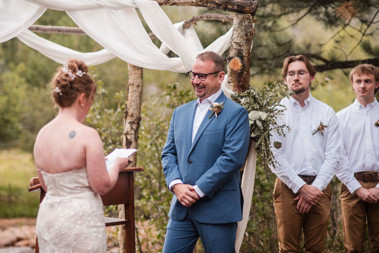 Groom in blue suit smiling during outdoor wedding ceremony under rustic arch with white draping and greenery. Vow exchange Estes park