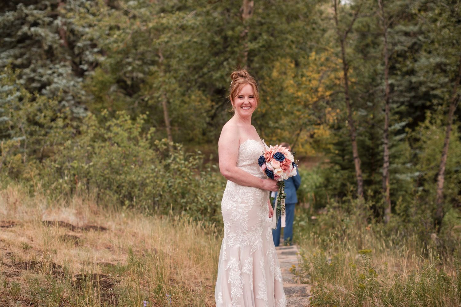 Bride in lace wedding gown holding colorful bouquet with blue ribbons, smiling outdoors in a forest setting. Looking back first look