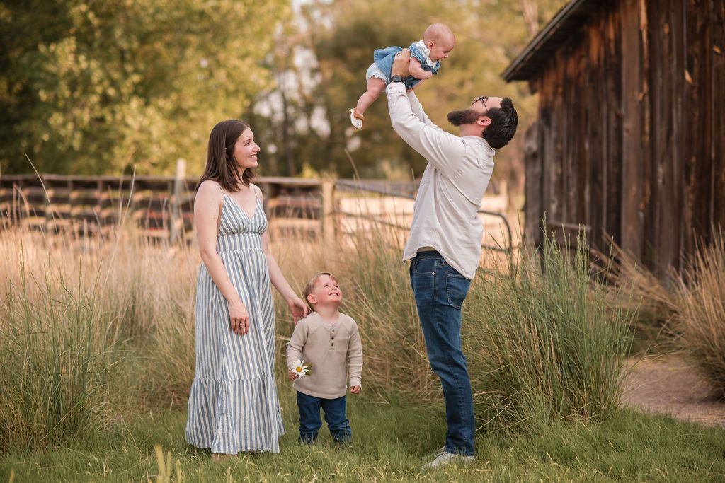 Family of four posing outdoors, father lifting baby in air, toddler and mother standing nearby, rustic barn background.  Mini sessions