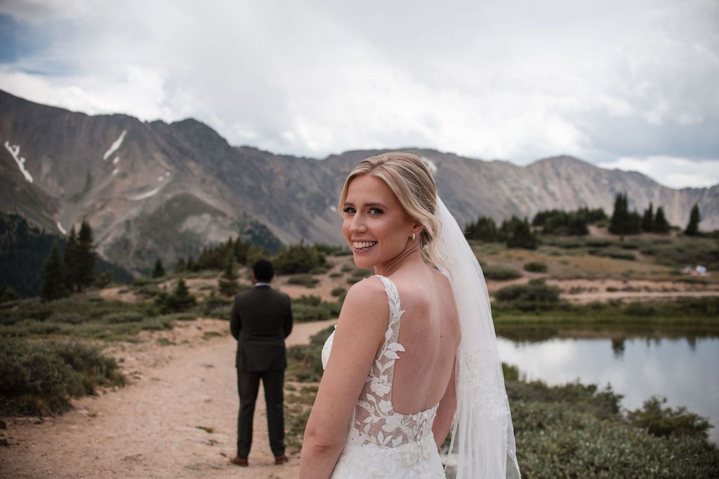 Bride in lace wedding gown and veil smiles over shoulder at mountain lake venue with groom standing ahead. First look Mountain Weddings