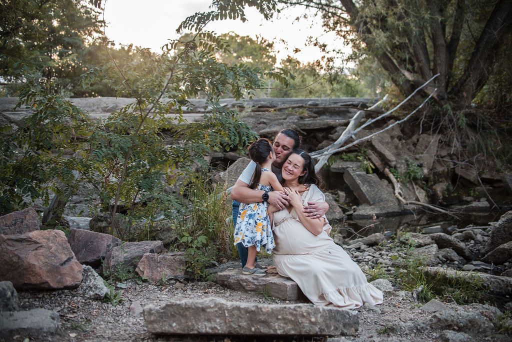 An interactive family portrait session showing a father hugging his wife and kissing his young daughter playfully on an outdoor trail.
