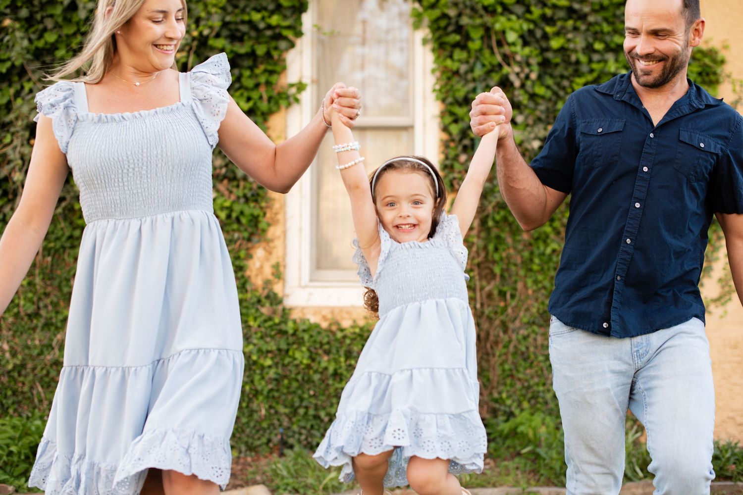 Happy family of three holding hands outdoors, parents swinging smiling girl in matching blue dresses on sunny day. Sandstone Ranch Longmont