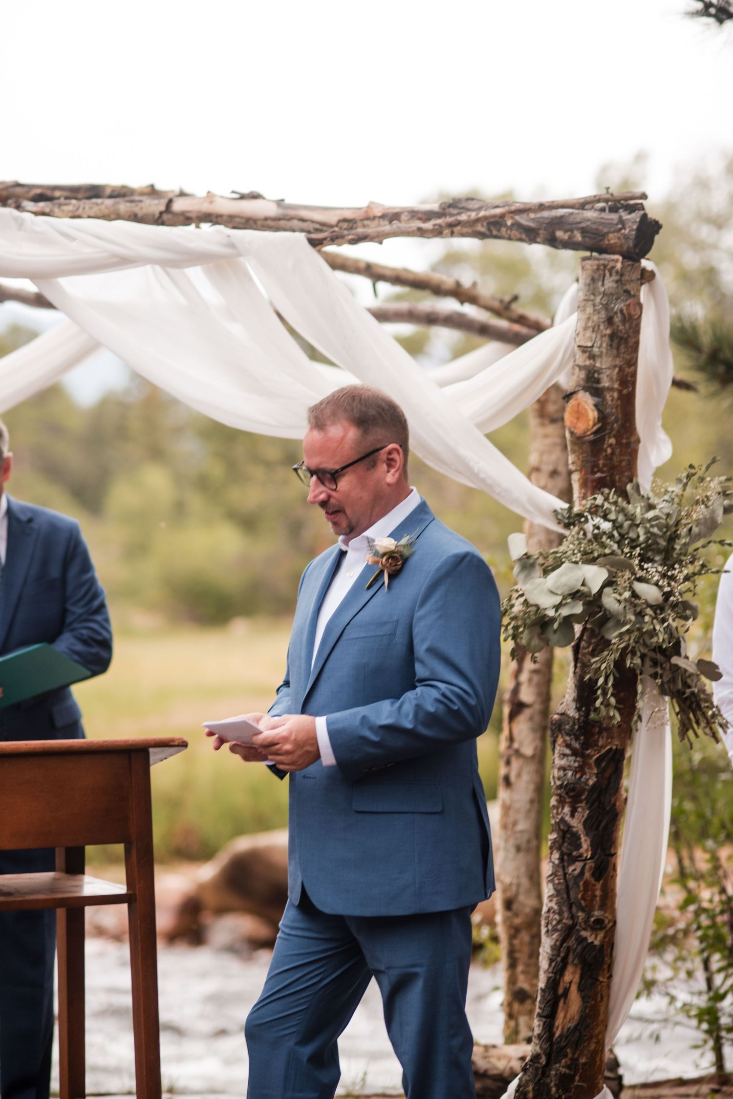 Groom in blue suit reading vows at rustic outdoor wedding ceremony with white draped arch and greenery.