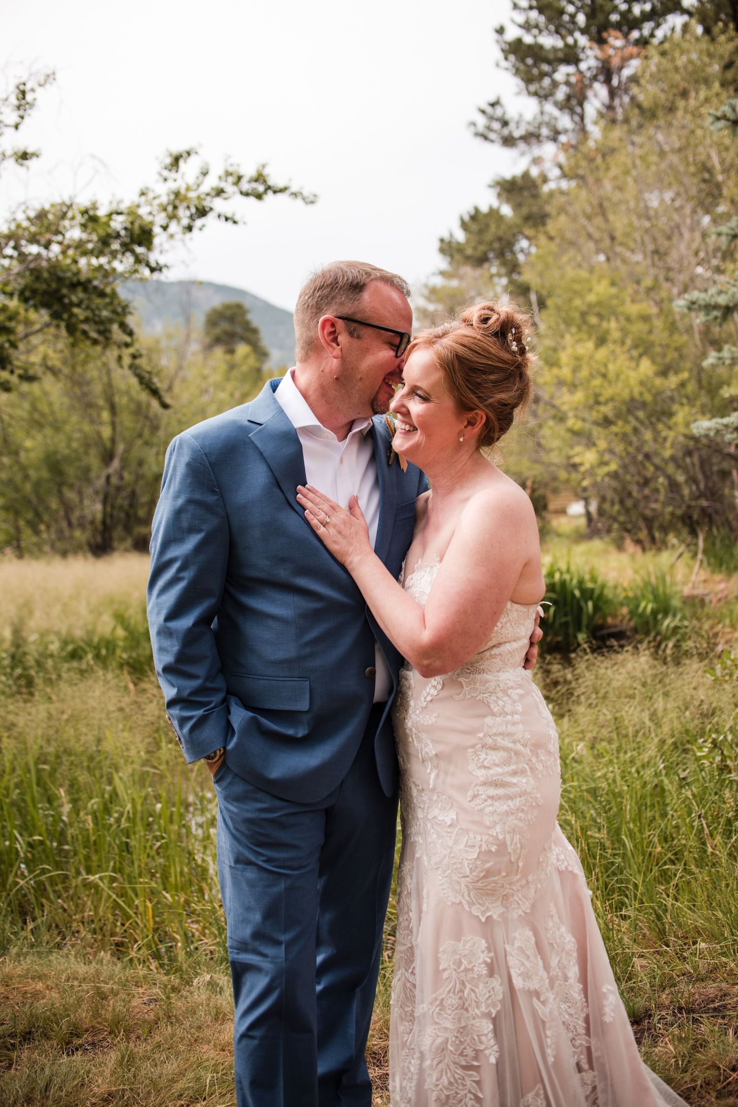 A bride and groom share a tender moment outdoors, with the groom in a blue suit whispering to his smiling bride in a lace gown.