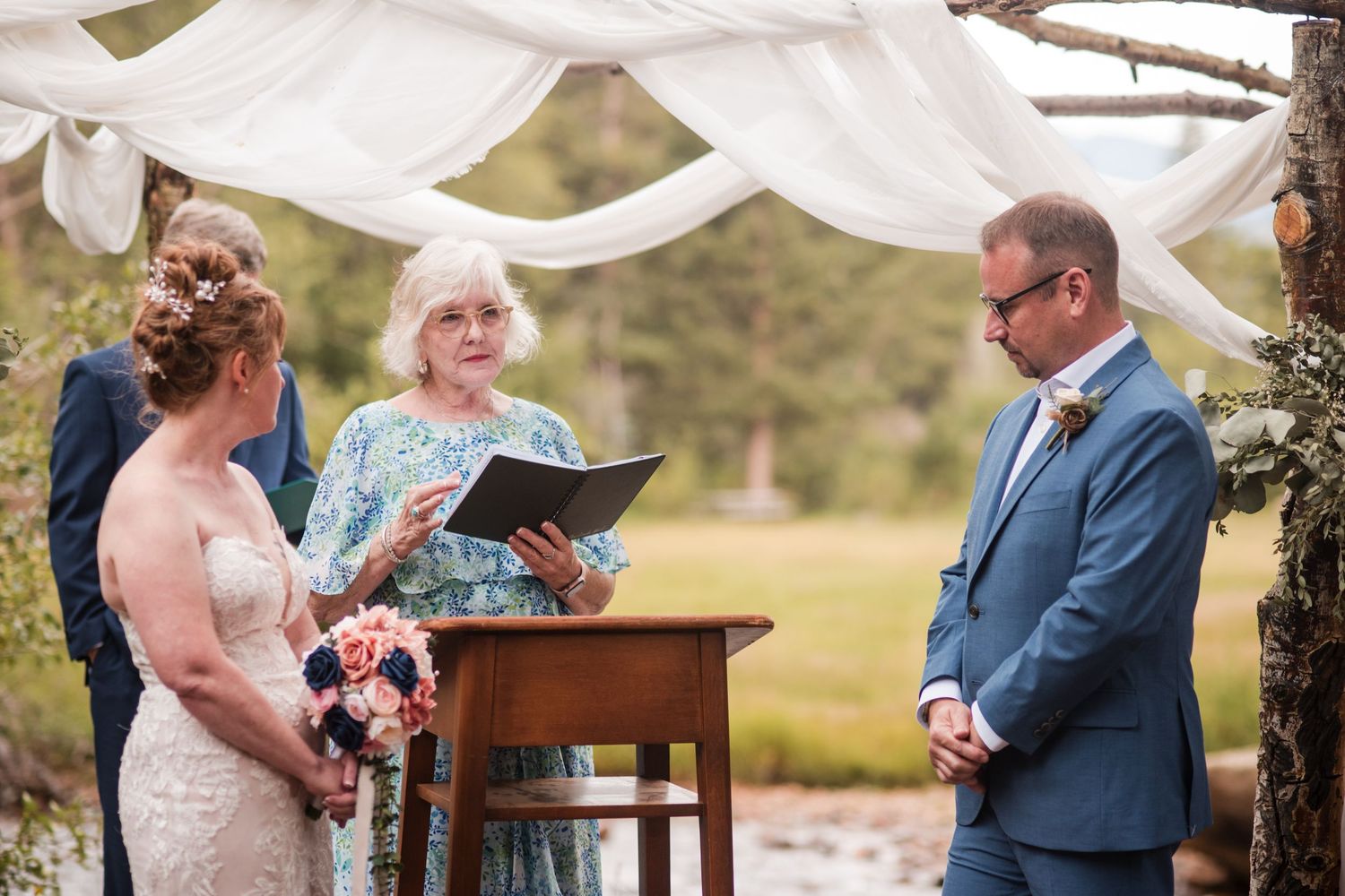 Outdoor wedding ceremony with bride, groom, and officiant under a white draped arbor in a scenic natural setting.
