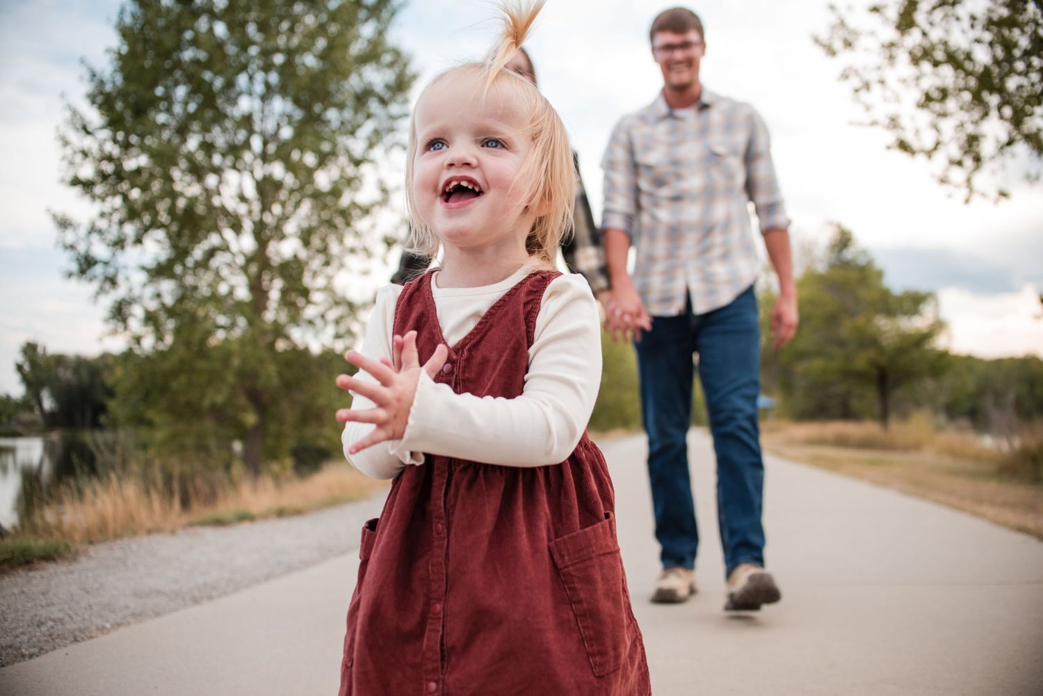 Joyful toddler girl in red dress laughing outdoors on a path with father walking behind her. Golden Ponds fall mini session