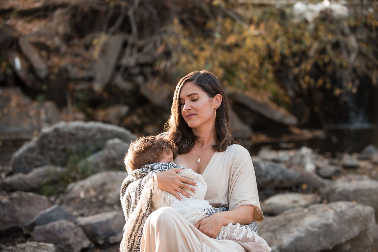 A mother tenderly holds her baby while sitting on rocks near a waterfall in soft natural light. Motherhood photo session at Golden Ponds