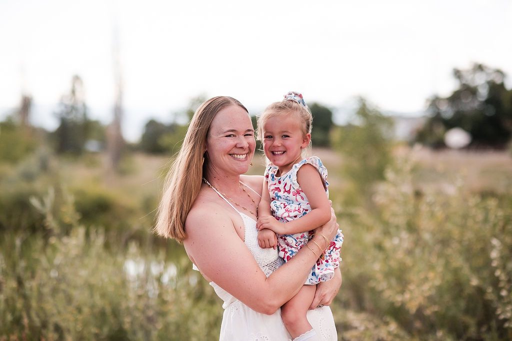 A smiling mother in a white dress holds her happy toddler girl in a floral outfit in a sunny meadow.Longmont and Boulder family photography