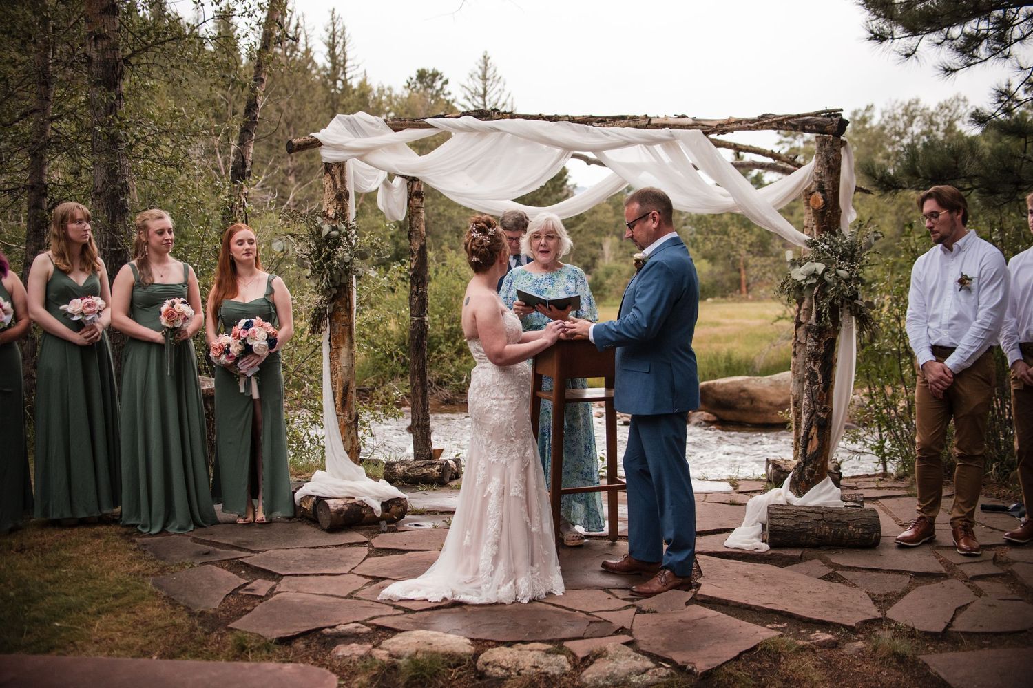 Outdoor wedding ceremony at rustic wooden arch with bride, groom, officiant, and bridesmaids in green dresses by a forest stream.