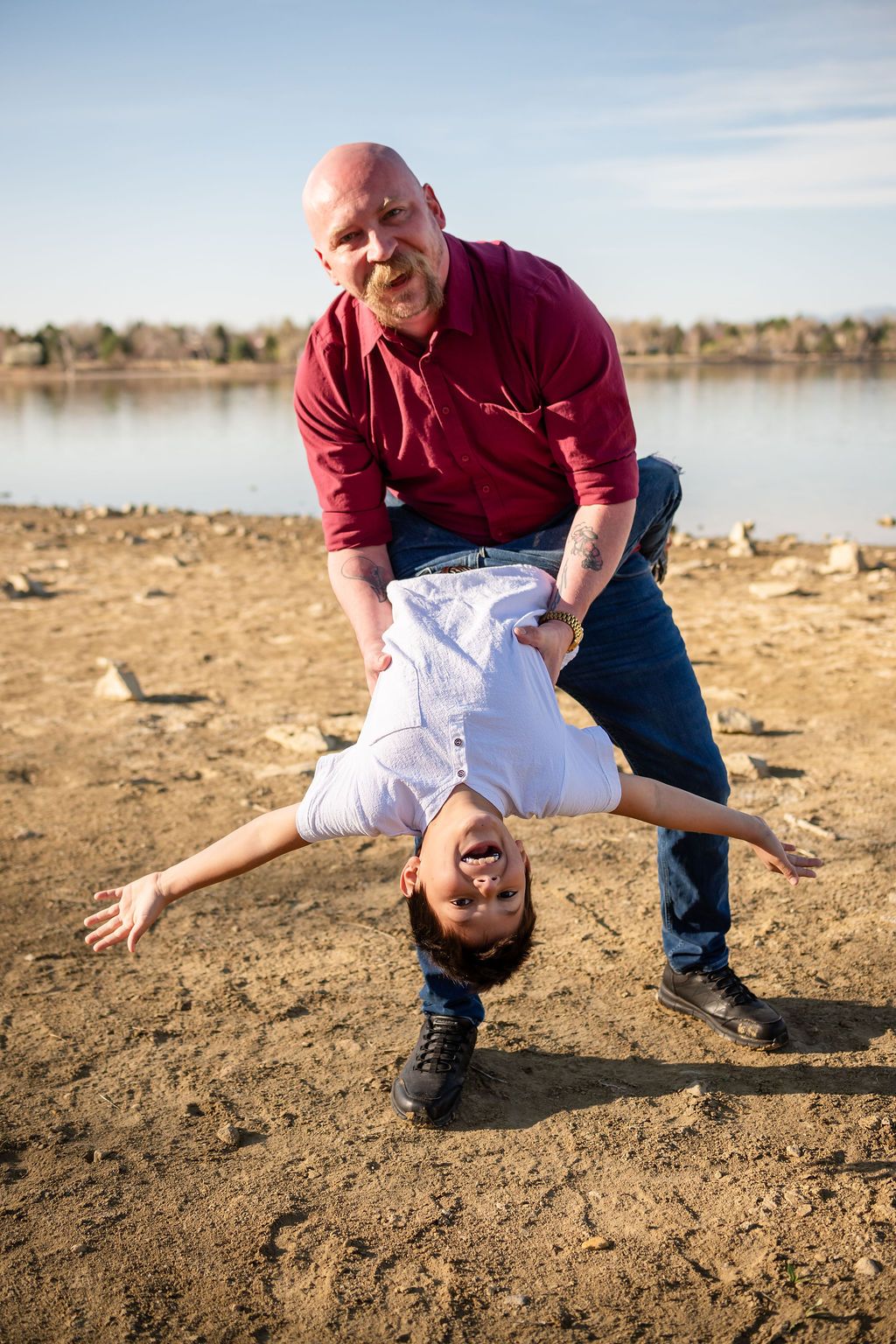 A father in a red shirt playfully holds a laughing child upside down on a sandy beach near a lake Family photographer McIntosh Lake Longmont