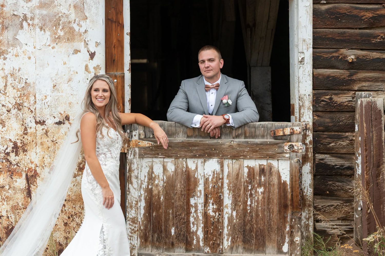 Bride in white gown and groom in gray suit pose at rustic barn door for wedding photos. Wedding at the Pines mountain wedding photography