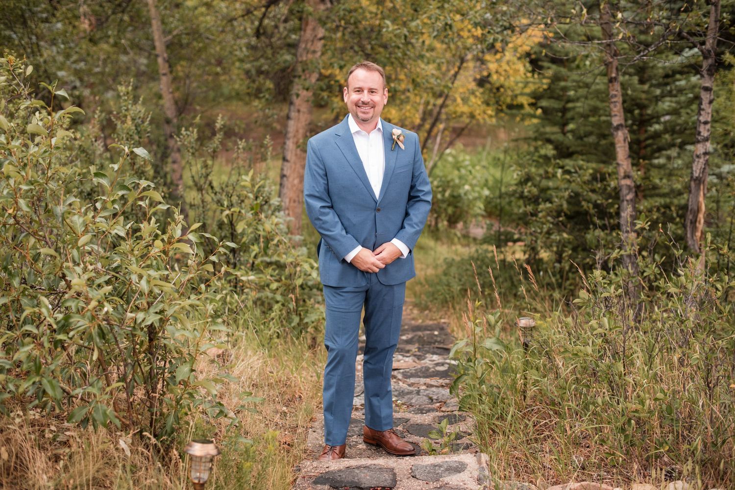 Groom in blue suit standing on stone path surrounded by lush green and autumn foliage at outdoor wedding. Waiting for his Bride first look
