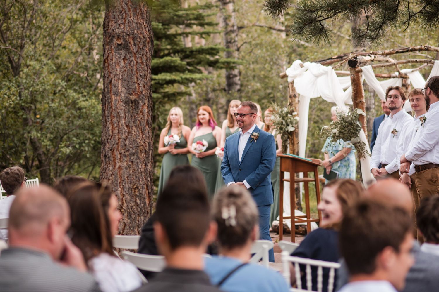 Groom stands at outdoor wedding altar surrounded by trees, with wedding party and seated guests in attendance. Looking at bride walking down