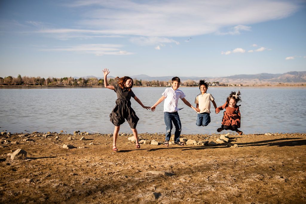 Four children holding hands and jumping joyfully on a rocky lakeshore under a bright blue sky. Family photography McIntosh Lake Longmont, CO