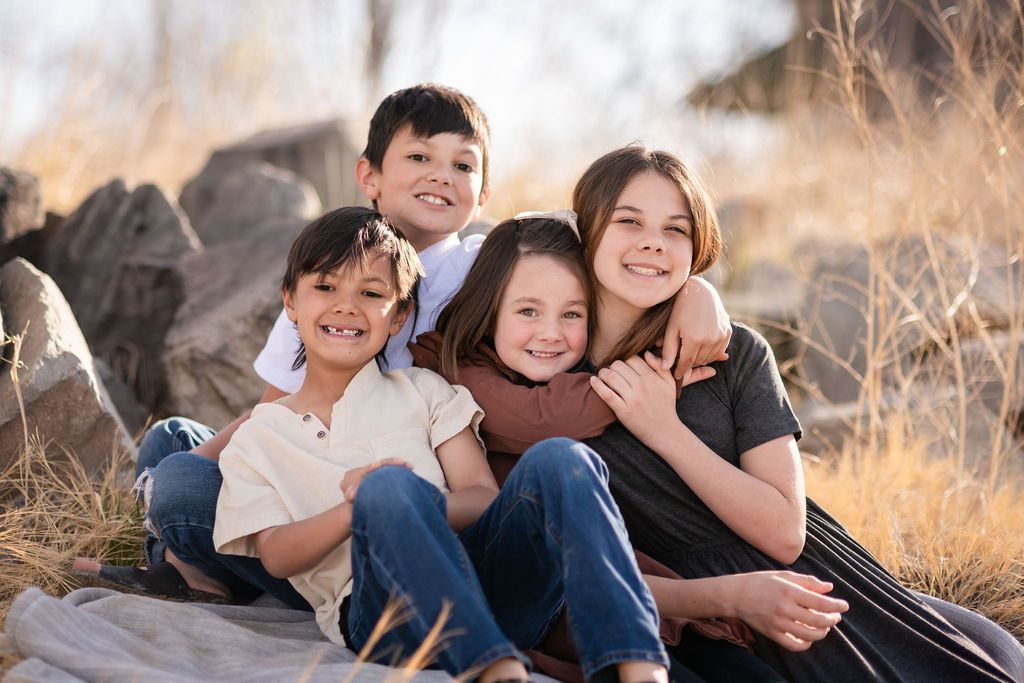 Four children smiling and hugging outdoors on a blanket with rocks and golden grass in the background. McIntosh Lake, Longmont Co