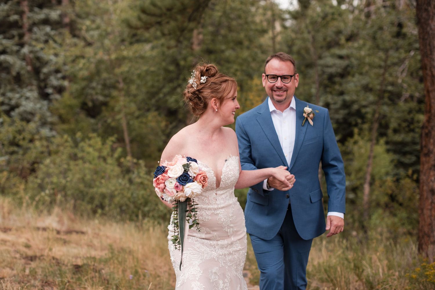 A bride and groom walk hand in hand through a forest meadow; she holds a bouquet as he wears a blue suit.-RiversongInnRetreat-Estes Park