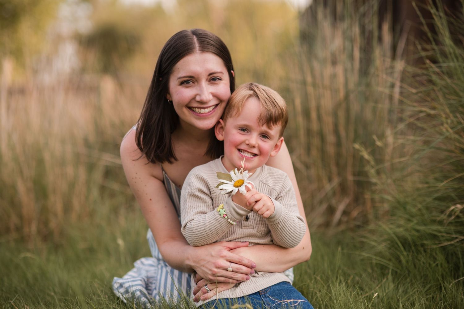 A smiling mother hugs her young son holding a daisy flower while sitting in a lush green field outdoors. at Sandstone Ranch Co for a photo