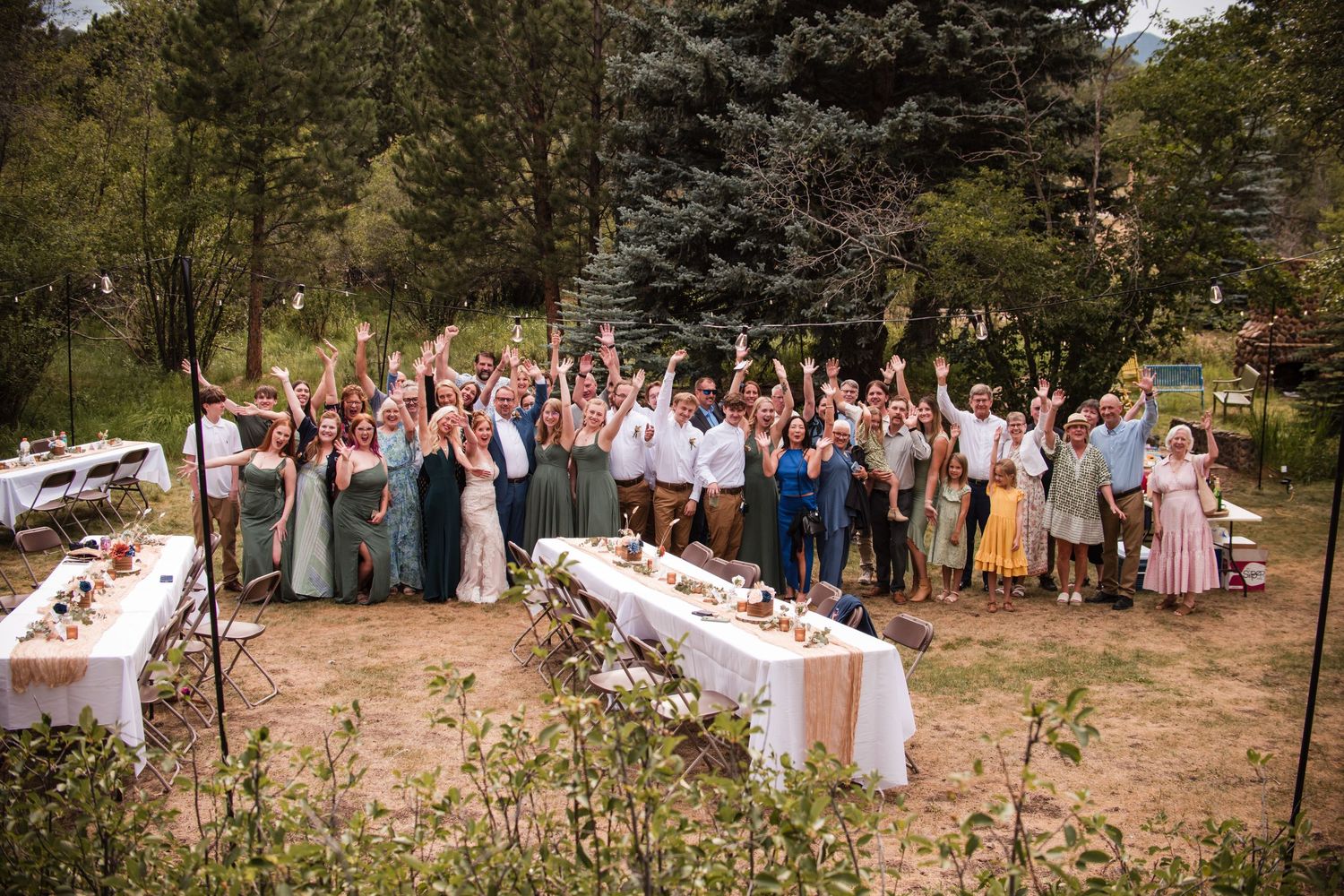 Large wedding party celebrating outdoors in a garden setting, raising hands joyfully near decorated tables. Group photo of all guests