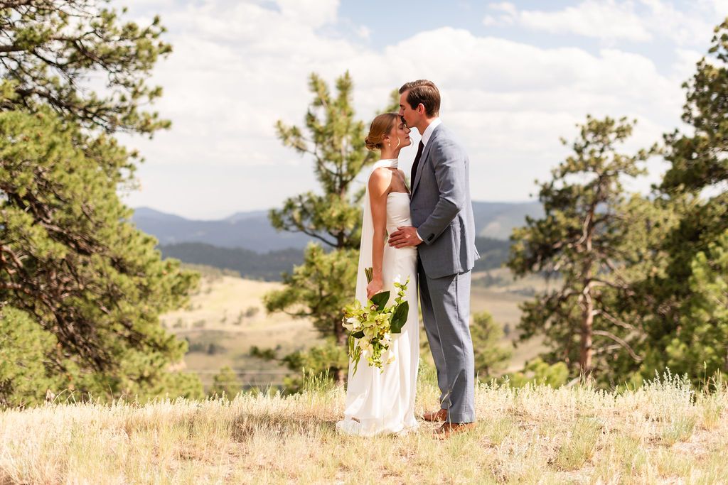 A bride and groom share a romantic moment in a scenic mountain meadow surrounded by pine trees on their wedding day. Golden Co Wedding