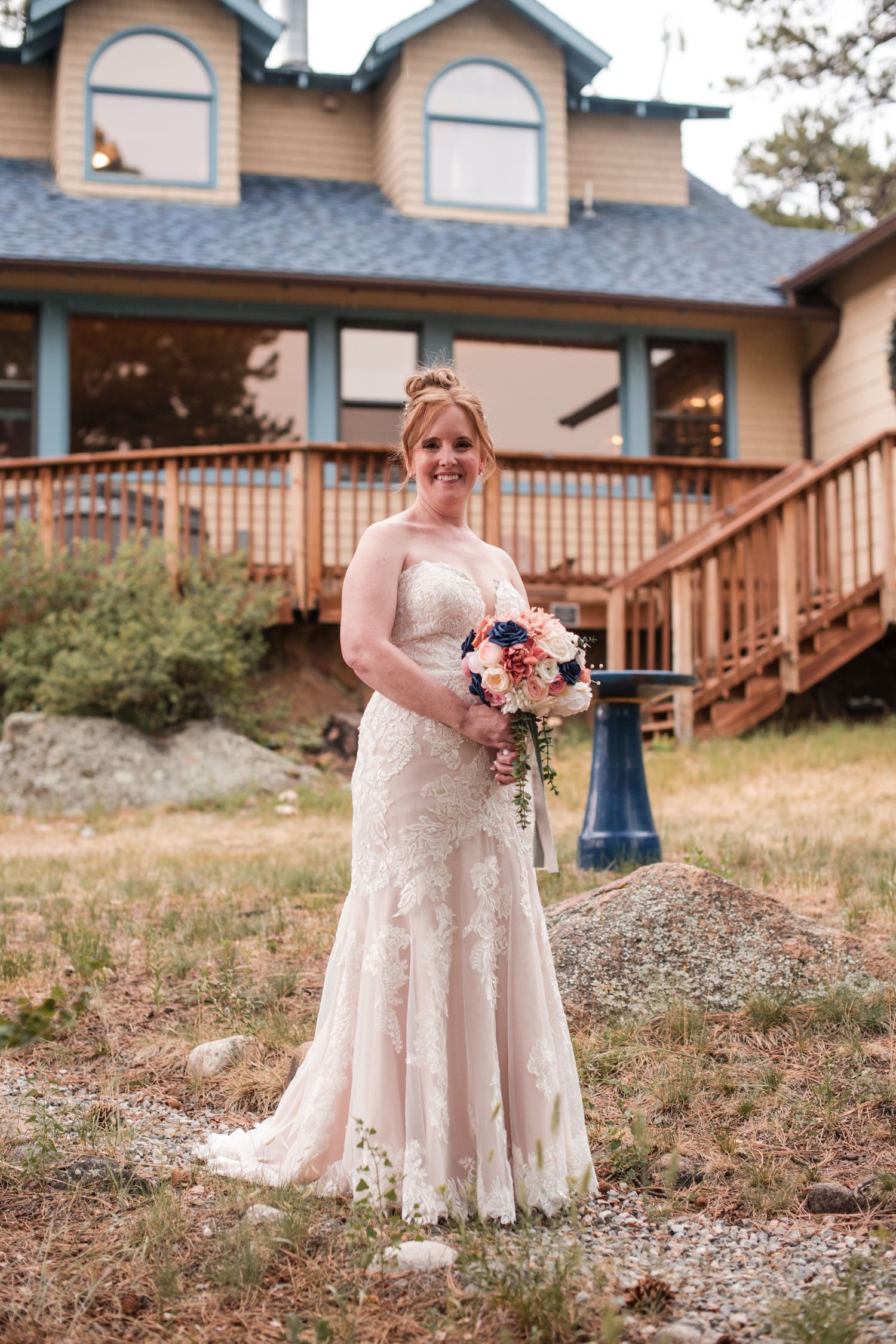 Bride in a lace wedding gown, holding a pink-and-navy bouquet, stands before a rustic mountain lodge with a wooden deck. Wedding Photography