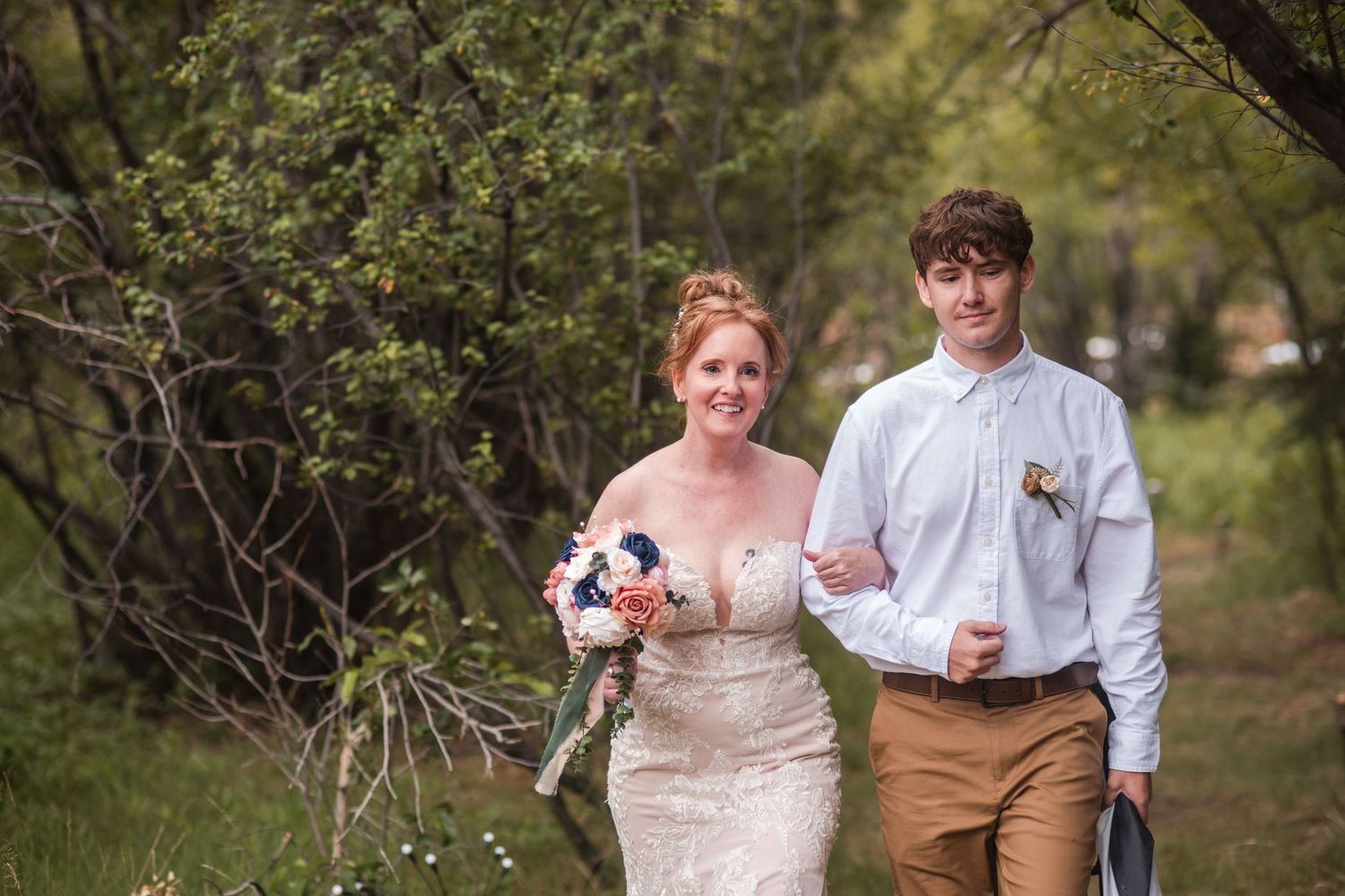 Bride in lace gown holding bouquet walks with son in white shirt through green wooded path on wedding day to altar. Estes Park wedding
