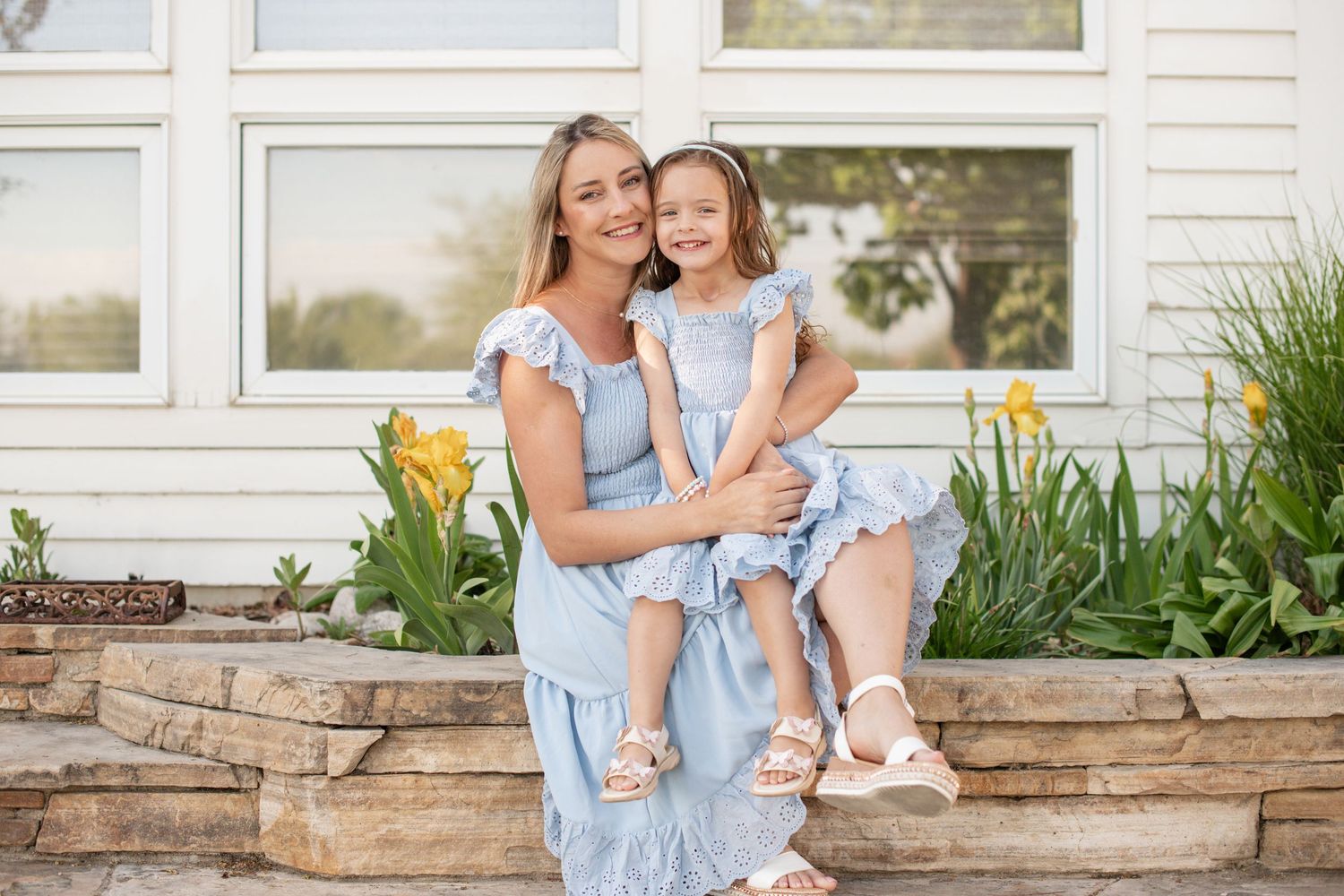 A mother and daughter in matching blue dresses smile together outdoors near yellow tulips and a stone wall.  Family photography, Longmont CO