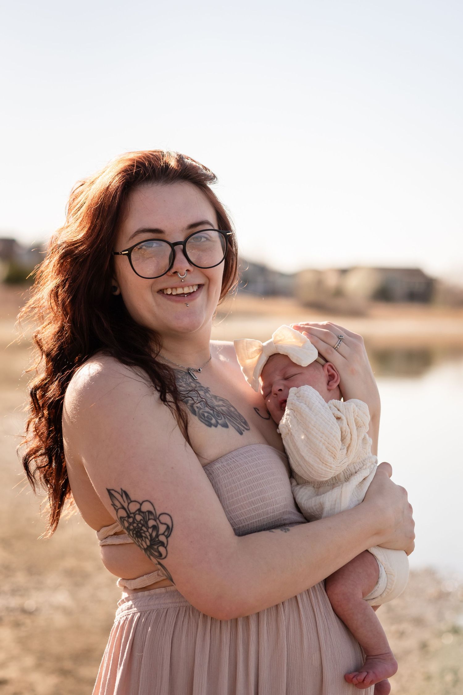A smiling young mother with red hair, glasses, and tattoos holds her newborn baby outdoors near a lake. Family session at McIntosh Lake