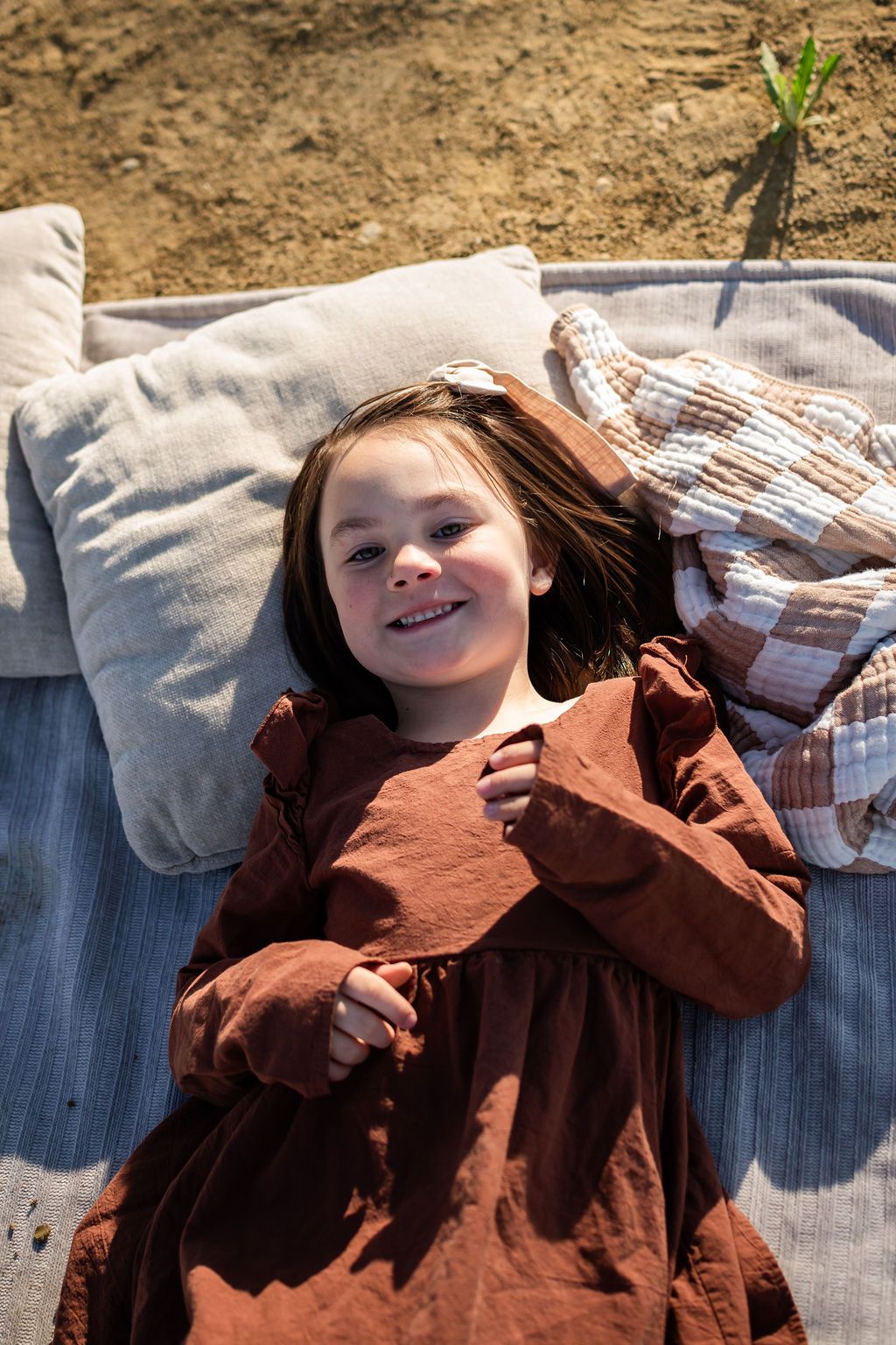 Smiling young girl in a rust-colored dress lying on a blanket with pillows outdoors in bright sunlight. At McIntosh Lake family photography