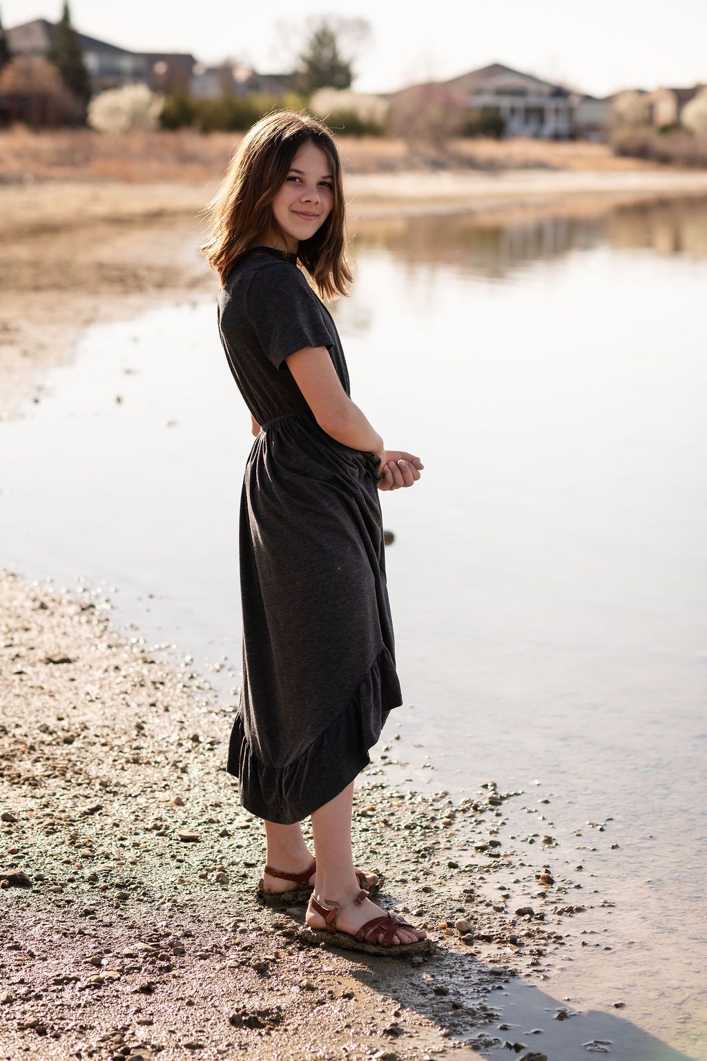Young woman in dark midi dress and sandals standing by a serene lake shore, smiling over her shoulder. Family photography McIntosh Lake