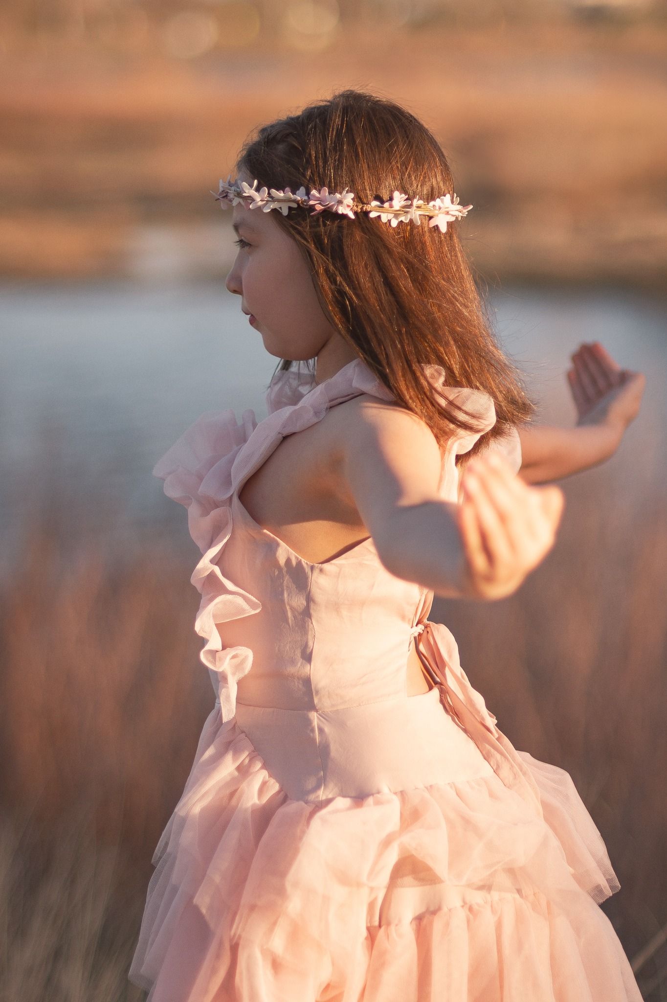 Young girl in a pink tulle dress and a floral crown dancing outdoors in golden-hour light. Longmont Child Fine Art Photographer Joyfolie