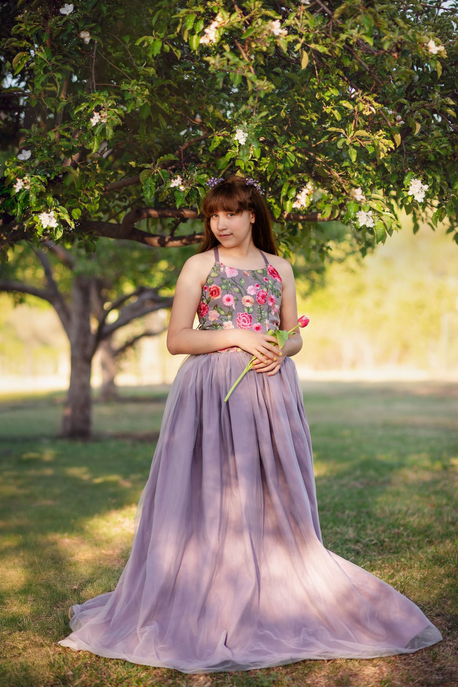 A young girl in a floral-embroidered lavender gown, holding a tulip under a blooming tree. Little Dreamers Couture Fine Art Photography
