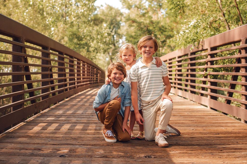Three young siblings laughing together while sitting on a rustic wooden footbridge at Golden Ponds Nature Area during a fall family mini ses