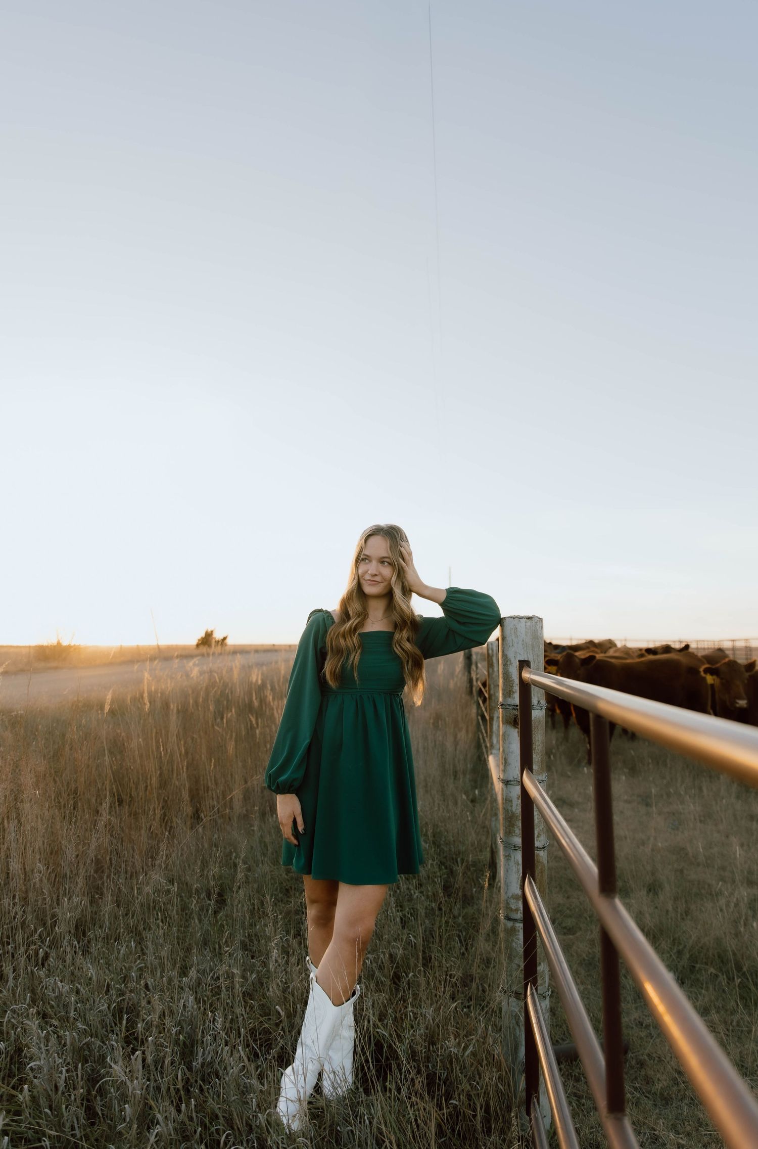 A woman in a green dress and white boots poses along a wooden fence at sunset in a rustic field setting.