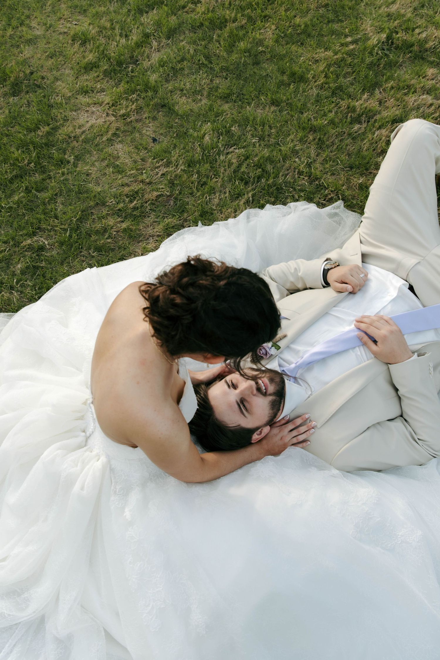 Bride in white gown leans over groom lying on grass in romantic outdoor wedding photo.