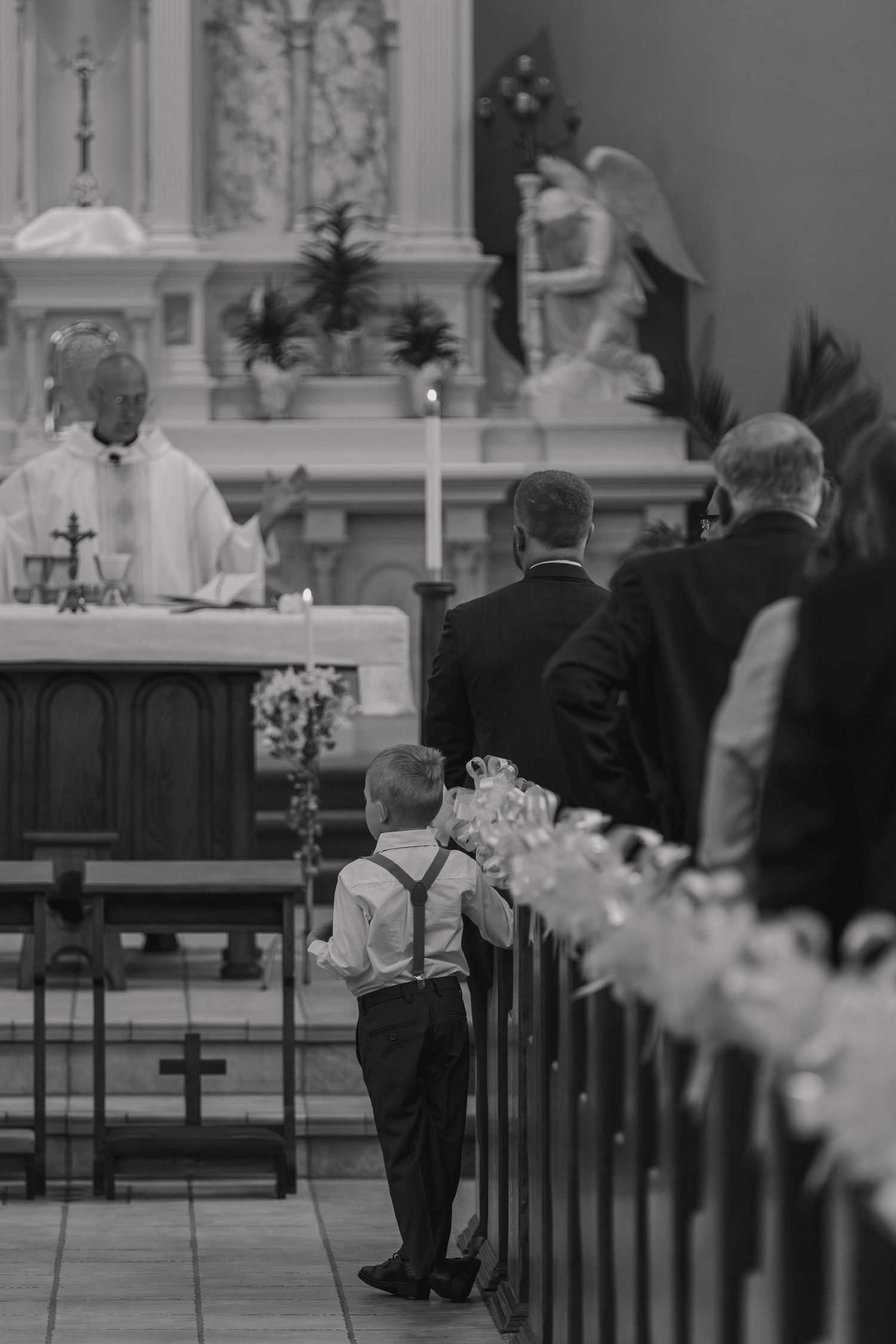 Black and white photo of a young boy in suspenders standing at church pew during a wedding ceremony with priest at altar.