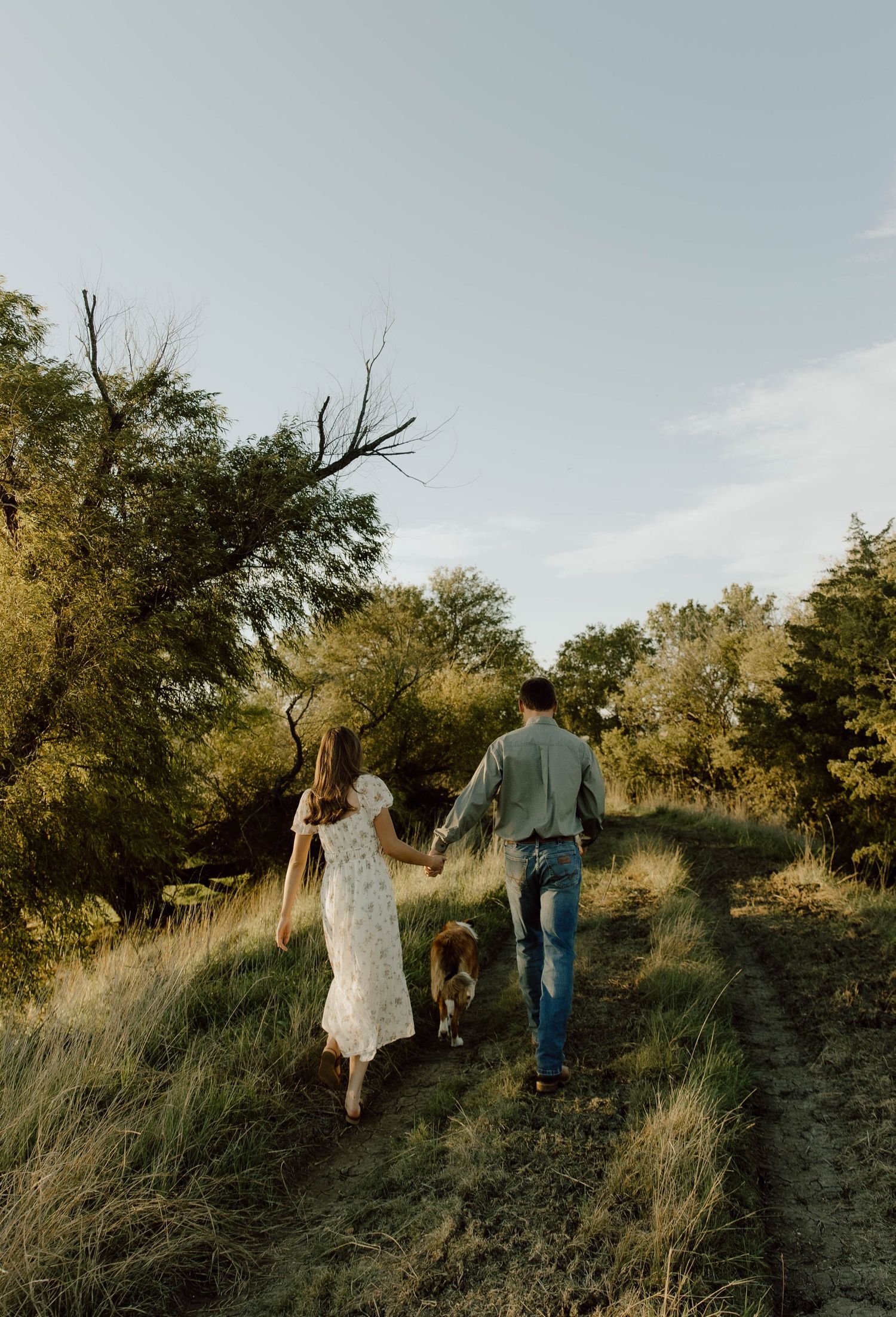 Couple walking up a grassy hillside path through trees at sunset in a romantic outdoor setting.
