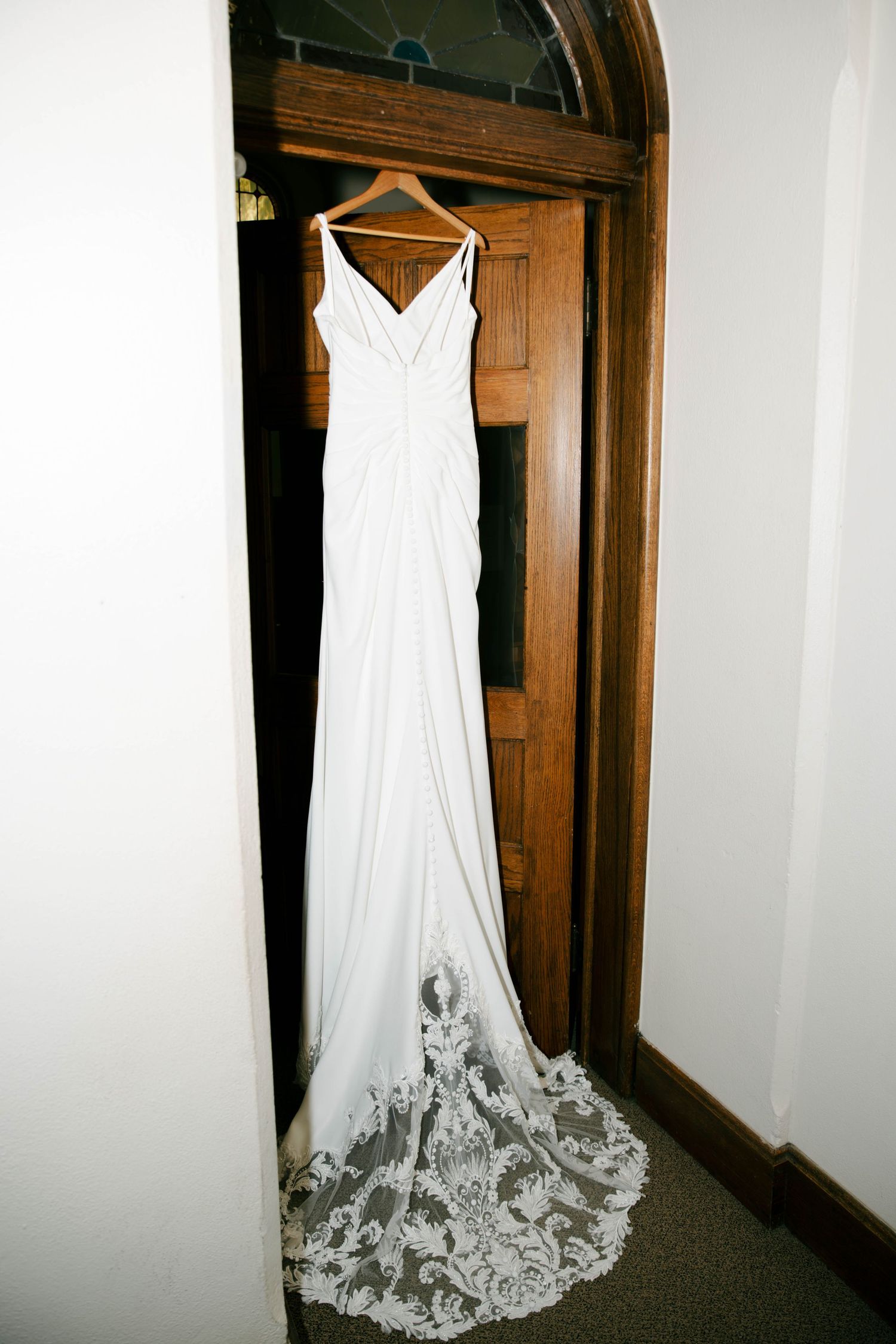 Elegant white wedding gown with lace train hanging on wooden door, featuring V-neck and sleek silhouette.
