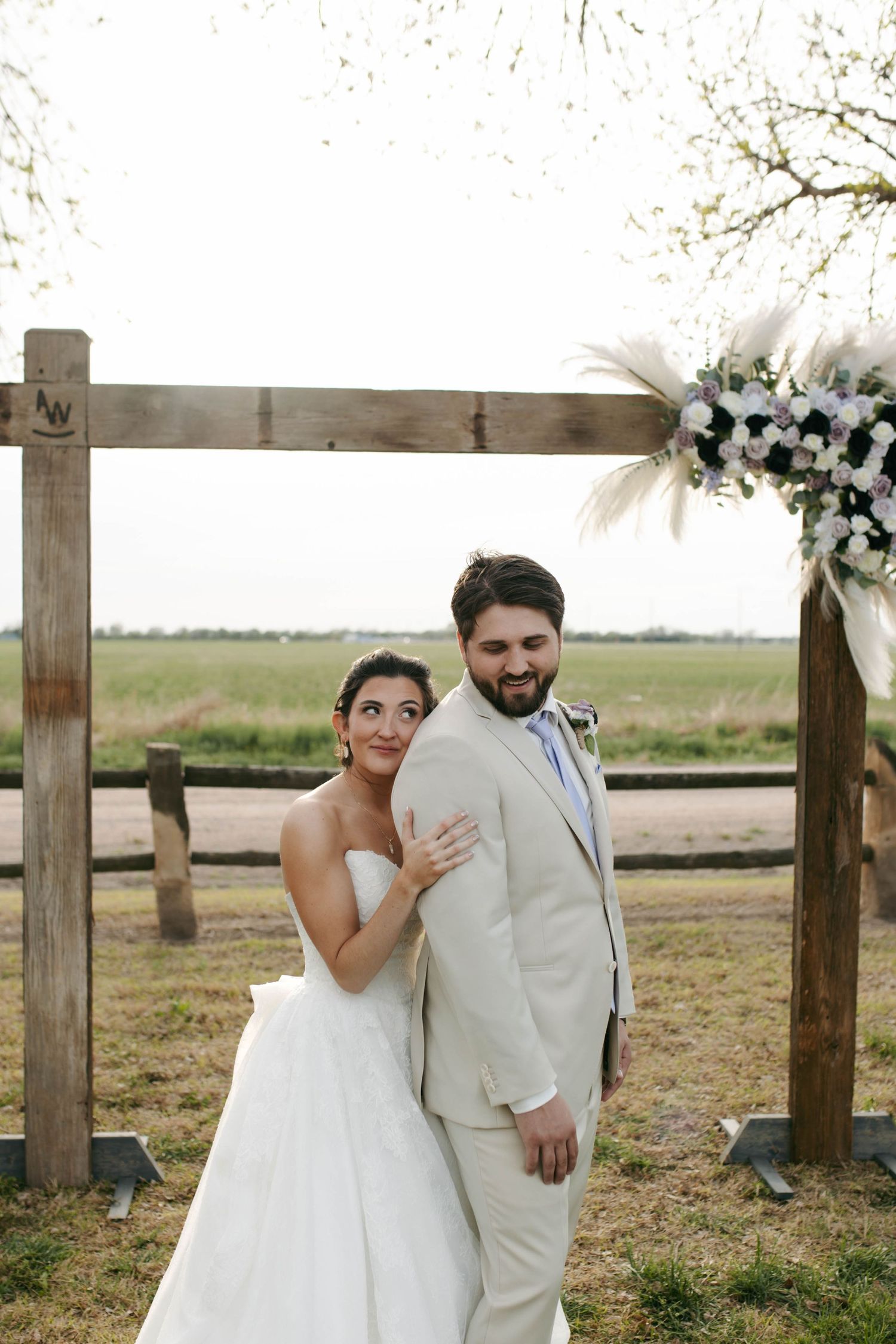 Bride and groom pose under rustic wooden arch with white floral arrangement at outdoor farm wedding.