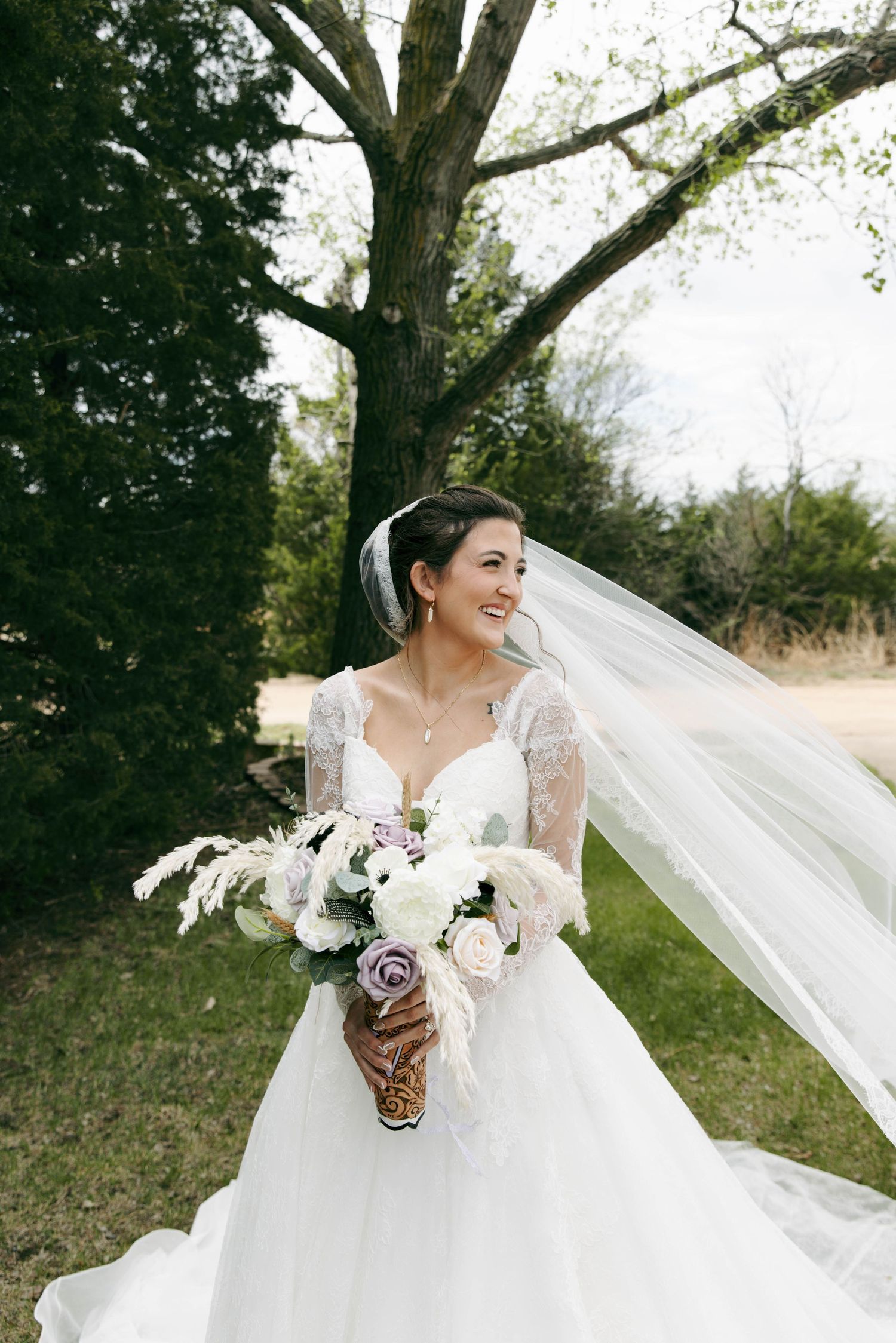 Smiling bride in lace wedding gown holds floral bouquet with veil flowing in breeze outdoors near large tree.