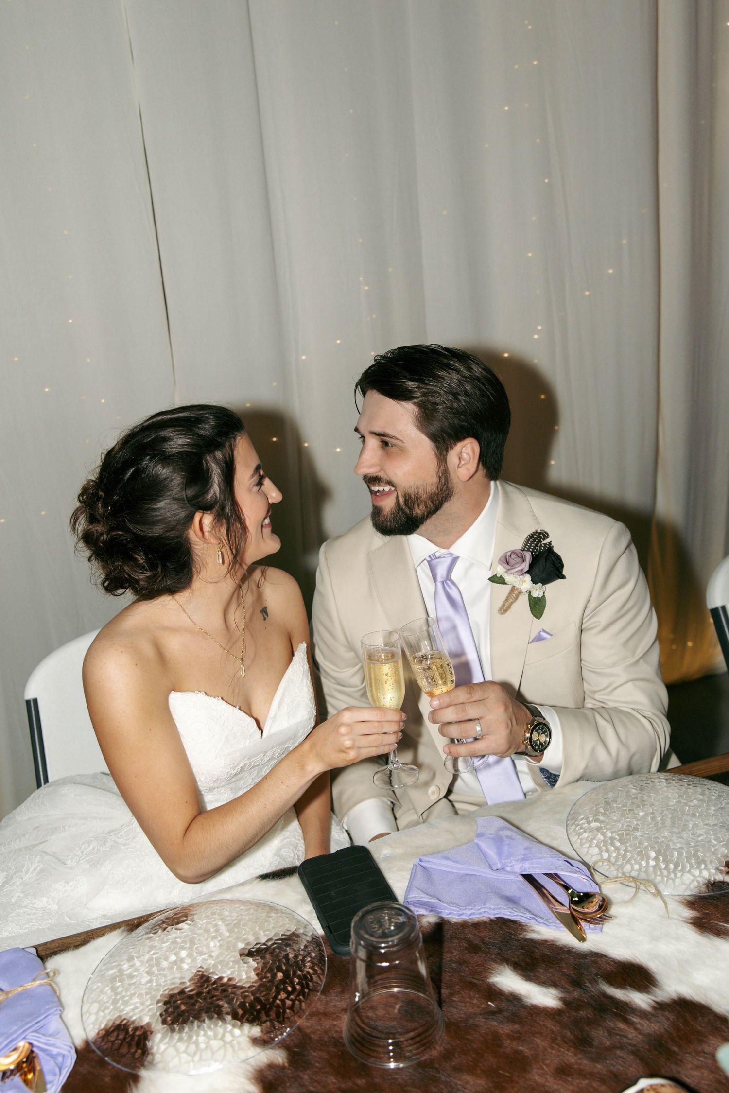 Bride and groom clinking champagne glasses at wedding reception table, smiling at each other in elegant attire.